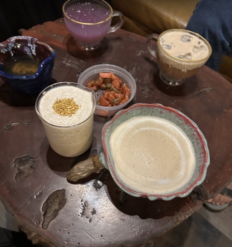 A spread of elixirs on a live-edge wood table — a lilac cup, an iced latte and a foam-topped glass dusted with pollen.
