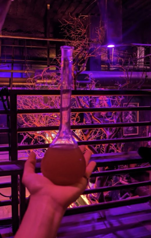 A hand raising a glass laboratory flask of amber elixir against violet light and bare branches.