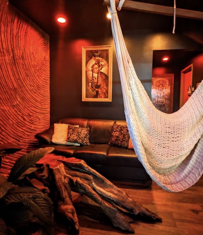 A single white hammock beside a leather couch and a driftwood table under warm amber light.